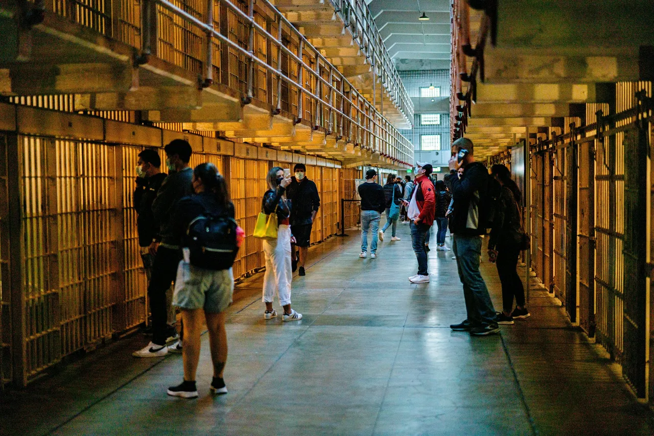 Visitatori con la mascherina lungo il corridoio di un vecchio carcere, fra due file di celle con le sbarre su più piani.