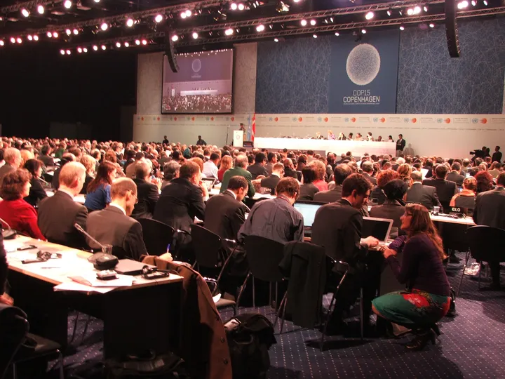 La sala plenaria della COP15 di Copenaghen durante la sessione di apertura: delegati seduti ai tavoli, sullo sfondo il palco con il logo della conferenza.