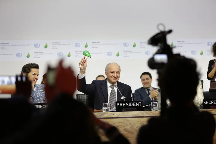 Il tavolo della presidenza della COP21 applaude dopo l'adozione dell'Accordo di Parigi, inquadrato tra le teste e le telecamere del pubblico.